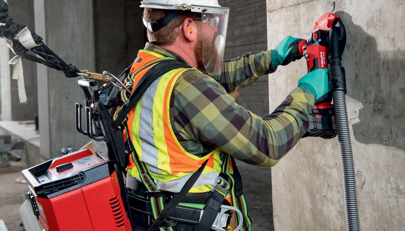 Een arbeider die boort in een betonnen muur draagt een snoerloze stofzuiger op zijn rug